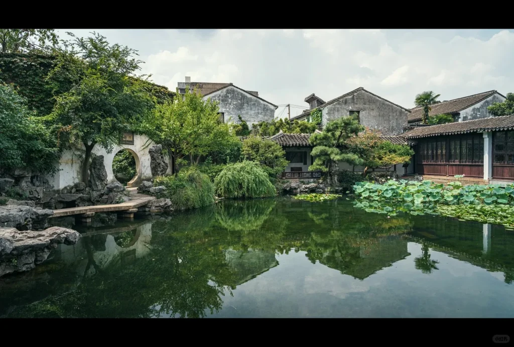 A classical Chinese garden featuring a tranquil pond with numerous green lotus leaves, reflecting traditional grey-tiled roof buildings and lush green trees. On the left, a white wall contains a circular moon gate, with a small stone bridge crossing a narrow section of the pond leading to it. Large decorative rocks are placed around the pond's edge.