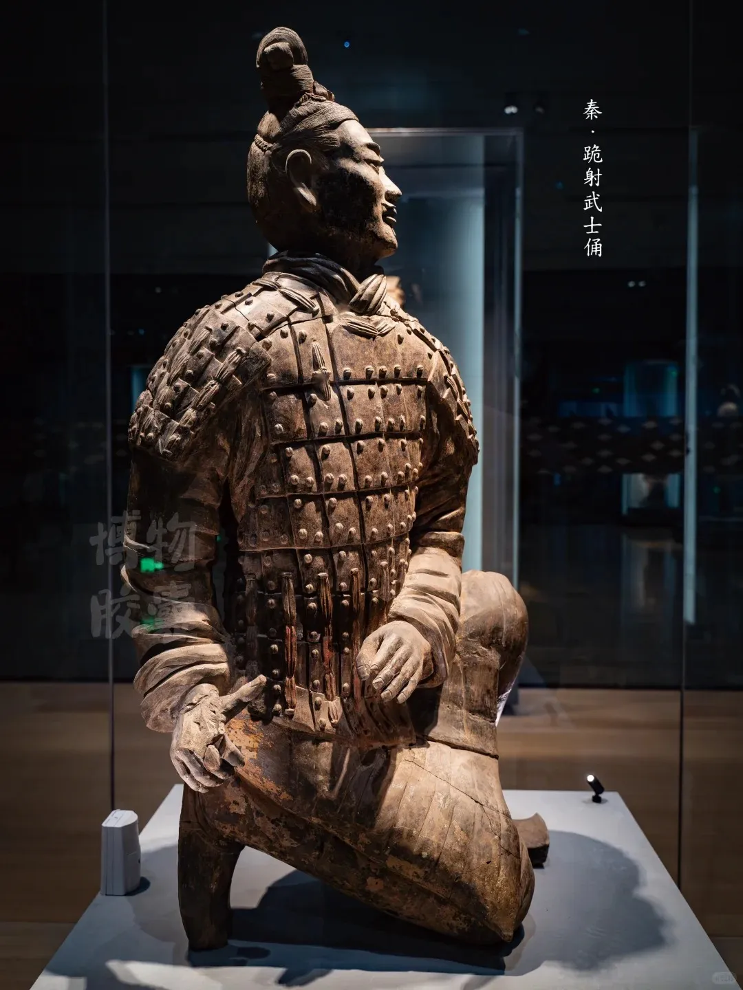 A detailed terracotta statue of a kneeling archer warrior. The ancient figure, made of clay, wears elaborate segmented armor with visible rivets and has its hair tied in a distinctive topknot. Its hands are positioned in front, ready to hold a bow, and its face is in profile, gazing slightly upwards. The statue is displayed in a dark museum setting, likely behind glass.