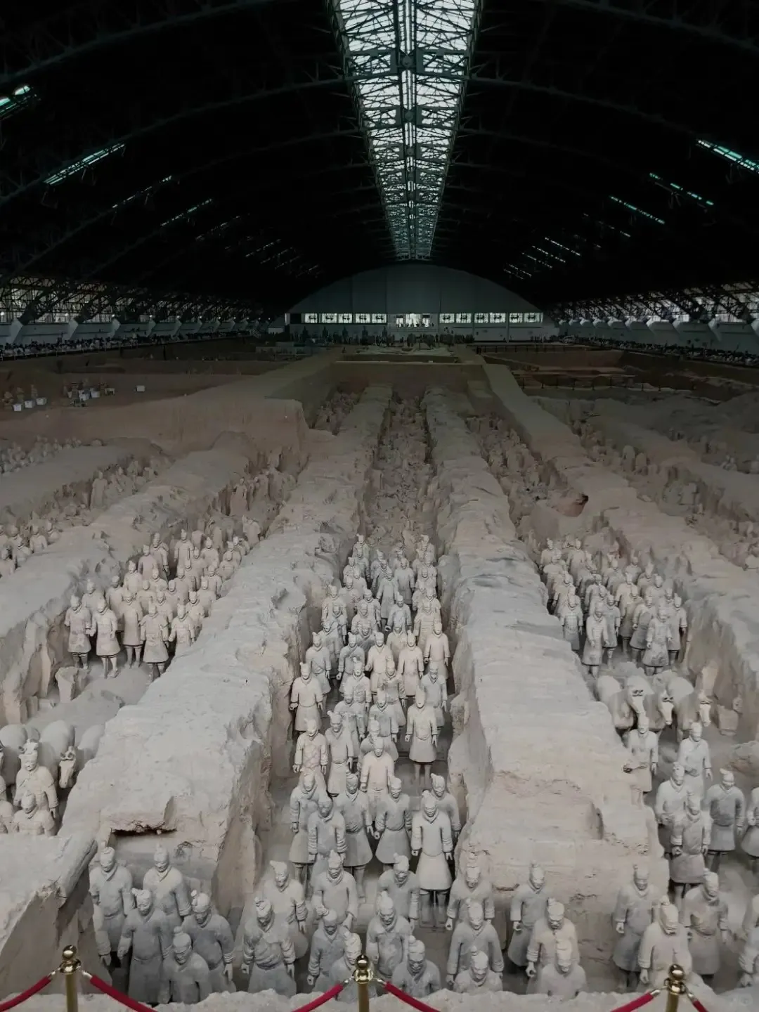 An elevated view of the expansive Pit 1 at the Terracotta Warriors museum, featuring hundreds of life-sized terracotta soldiers arranged in long rows within excavated trenches. The immense hall is covered by a high, dark, arched roof with skylights.