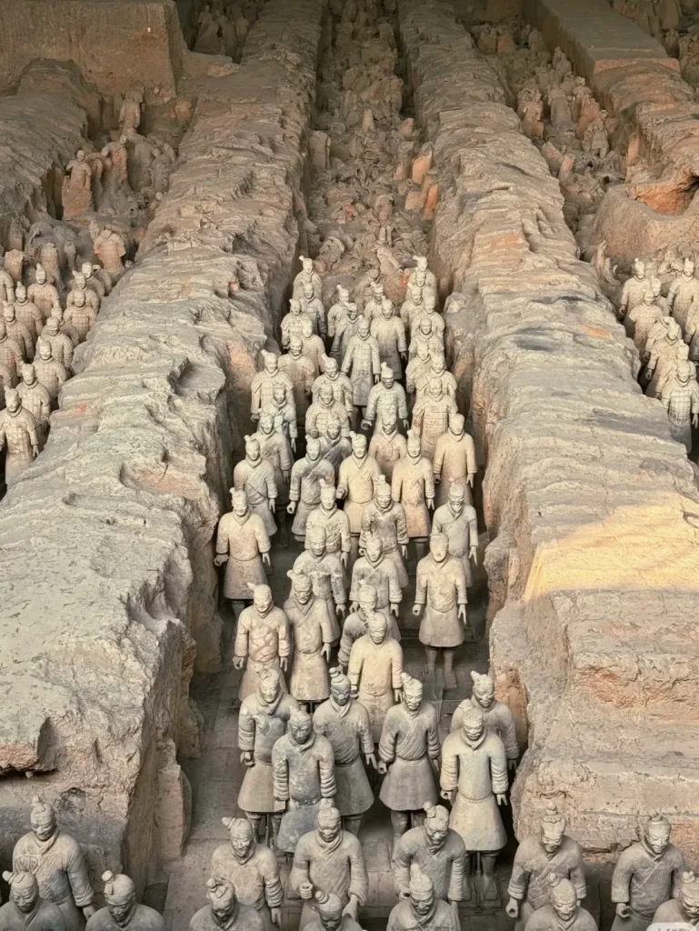 Rows of ancient Terracotta Warriors standing in formation within an excavated pit, with some warriors partially unearthed and others fully restored, at the historical site in Xi'an, China.
