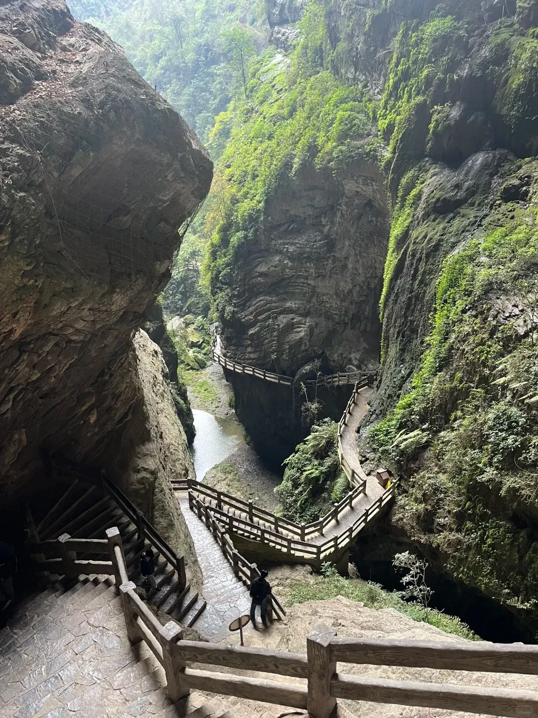 A deep, narrow gorge with towering, moss-covered rock walls. A series of wooden walkways with railings are built into the cliffside, winding alongside a shallow stream at the bottom. In the foreground, wide stone steps with wooden railings descend, with two individuals visible on them. More walkways curve into the distance, surrounded by lush green foliage.