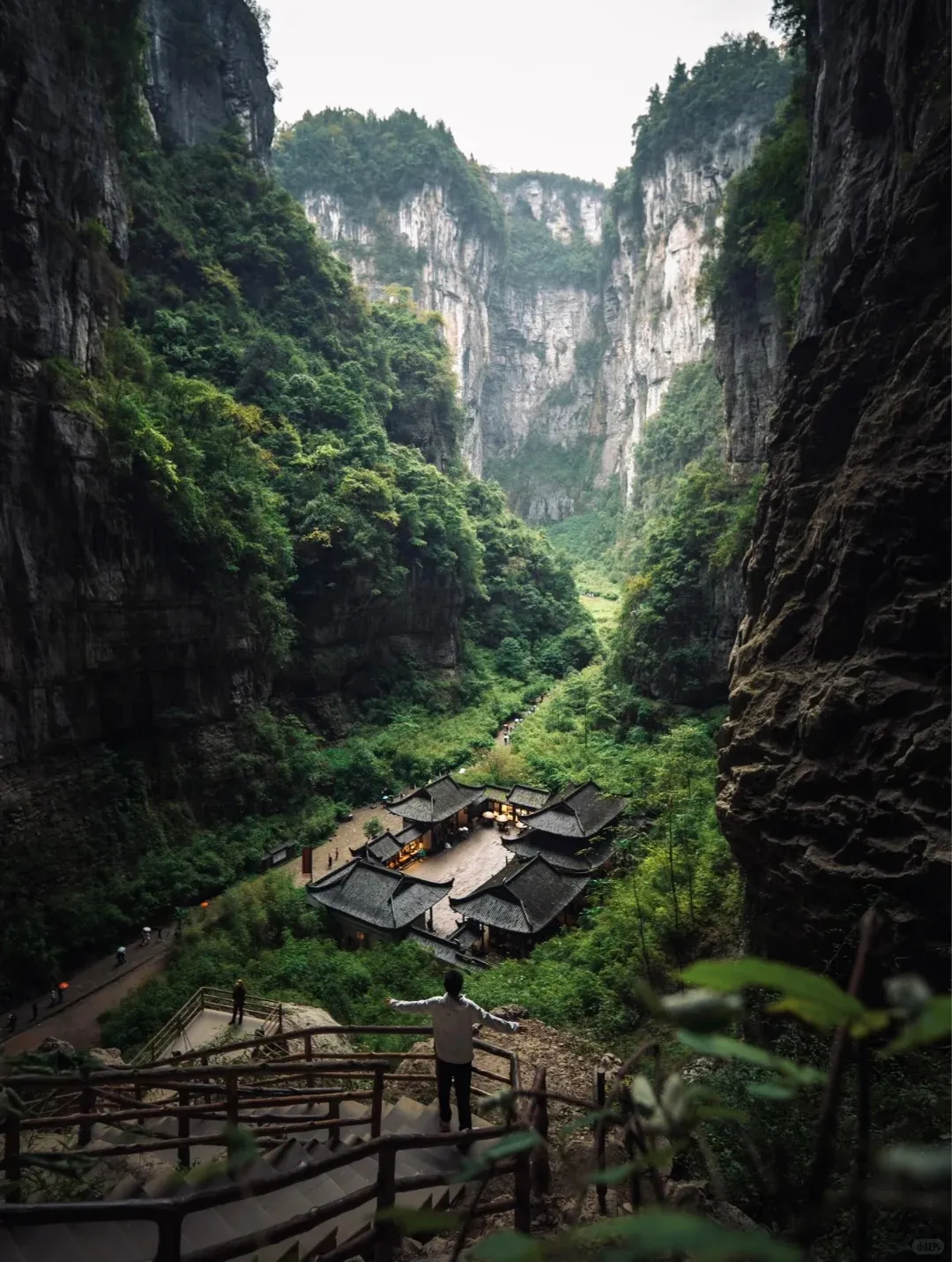 A breathtaking panoramic view of a deep, lush green karst canyon with towering limestone cliffs on either side. In the valley floor, a cluster of traditional Chinese-style buildings with dark tiled roofs is nestled amidst dense green foliage. A person stands in the foreground on a wooden staircase, arms outstretched, looking down into the expansive canyon.