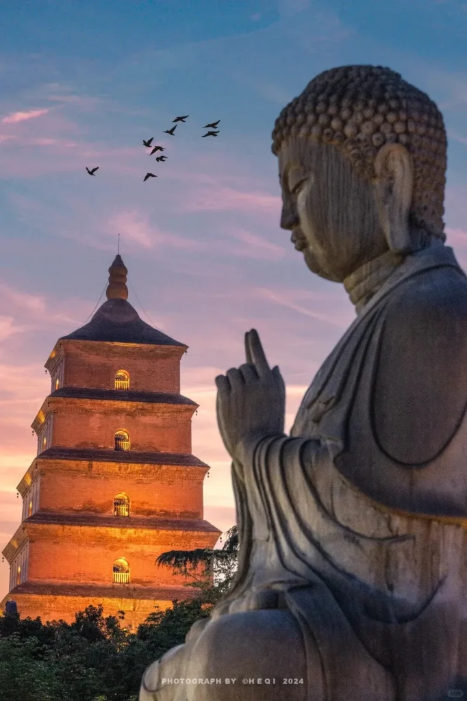A large stone Buddha statue in profile, with a hand raised, occupies the right side of the frame, slightly out of focus. In the left background, an illuminated, orange-red brick pagoda with multiple tiers and arched windows stands against a purple and blue dusk sky. A flock of birds flies in the upper left, and dark trees are visible at the pagoda's base.