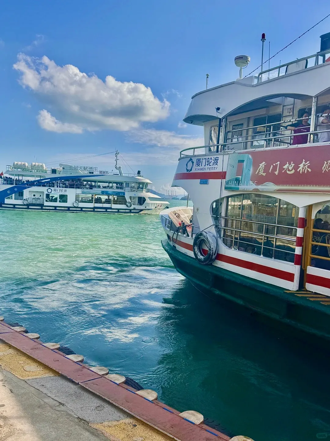 Two Xiamen ferries on turquoise water. The closer ferry is white with red stripes, docked at a concrete pier edge, displaying 'Xiamen Ferry' and 'Xiamen Landmark Ferry'. A second white and blue ferry is visible further in the background. The sky is blue with white clouds.