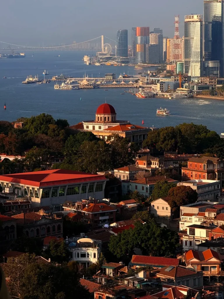 Panoramic view of Gulangyu Island's historic architecture, including a white building with a red dome, surrounded by trees in the foreground. A body of water with various ferries and boats separates it from the modern Xiamen city skyline in the background, which features numerous skyscrapers, a suspension bridge, and a Ferris wheel under a clear sky.