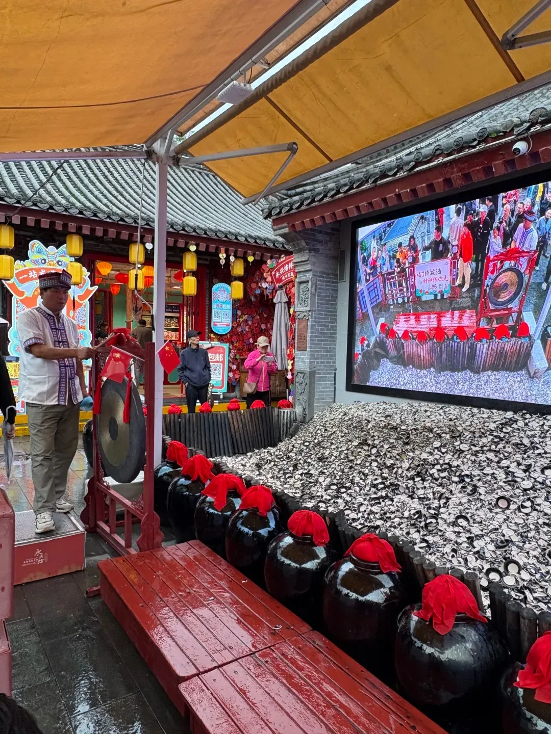 A man in a white tunic and patterned hat stands next to a large black gong on a red wooden frame. In the foreground, several large black ceramic jars with red cloths on top line a red wooden platform. Behind them, a large mound of shattered white and black ceramic bowls is piled up. A large outdoor digital screen in the background displays a similar festive scene with people.