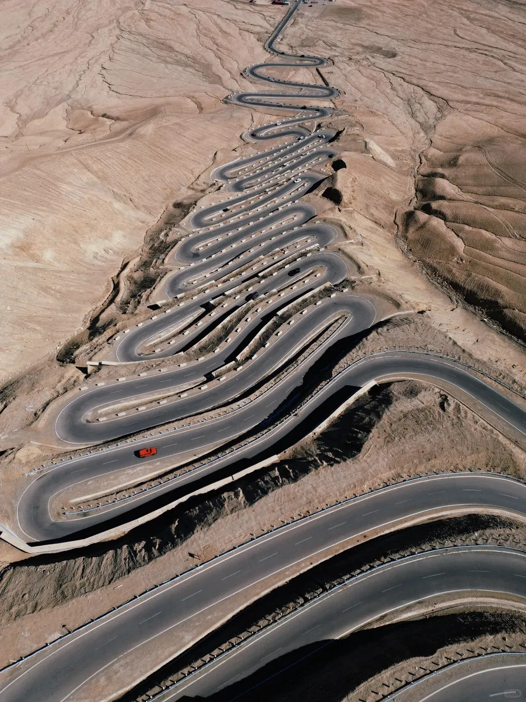 Aerial view of a paved mountain road with numerous tight switchbacks zigzagging down a barren, reddish-brown hillside. A single red car is visible on a lower curve, with smaller vehicles scattered along the upper sections of the road.