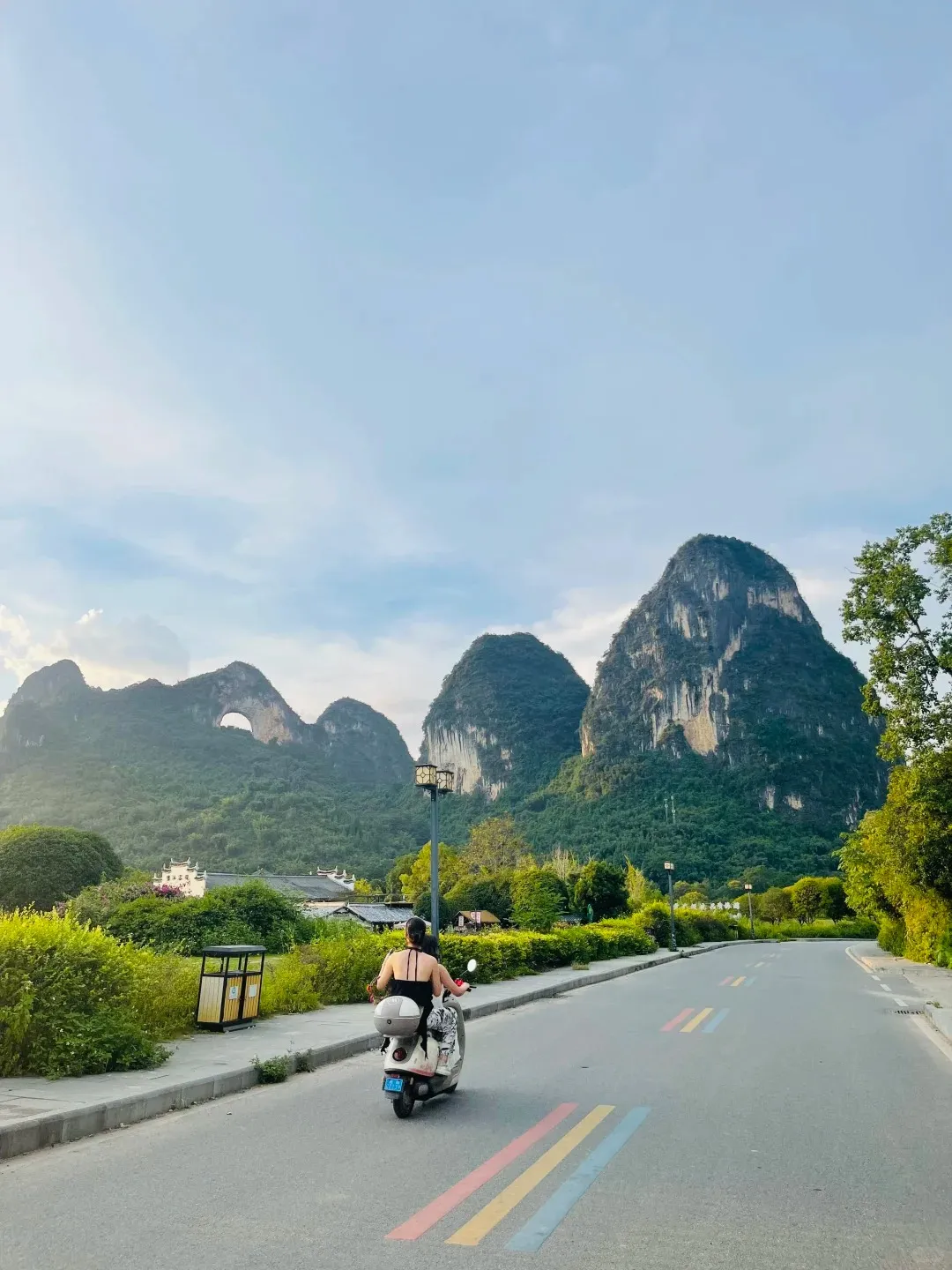 A person in a black top rides a white scooter away from the viewer on a paved road with colorful red, yellow, and blue stripes. The road is bordered by green bushes and a street lamp. In the background, tall, forested karst mountains rise, including one with a distinctive natural arch (Moon Hill), under a light blue sky.