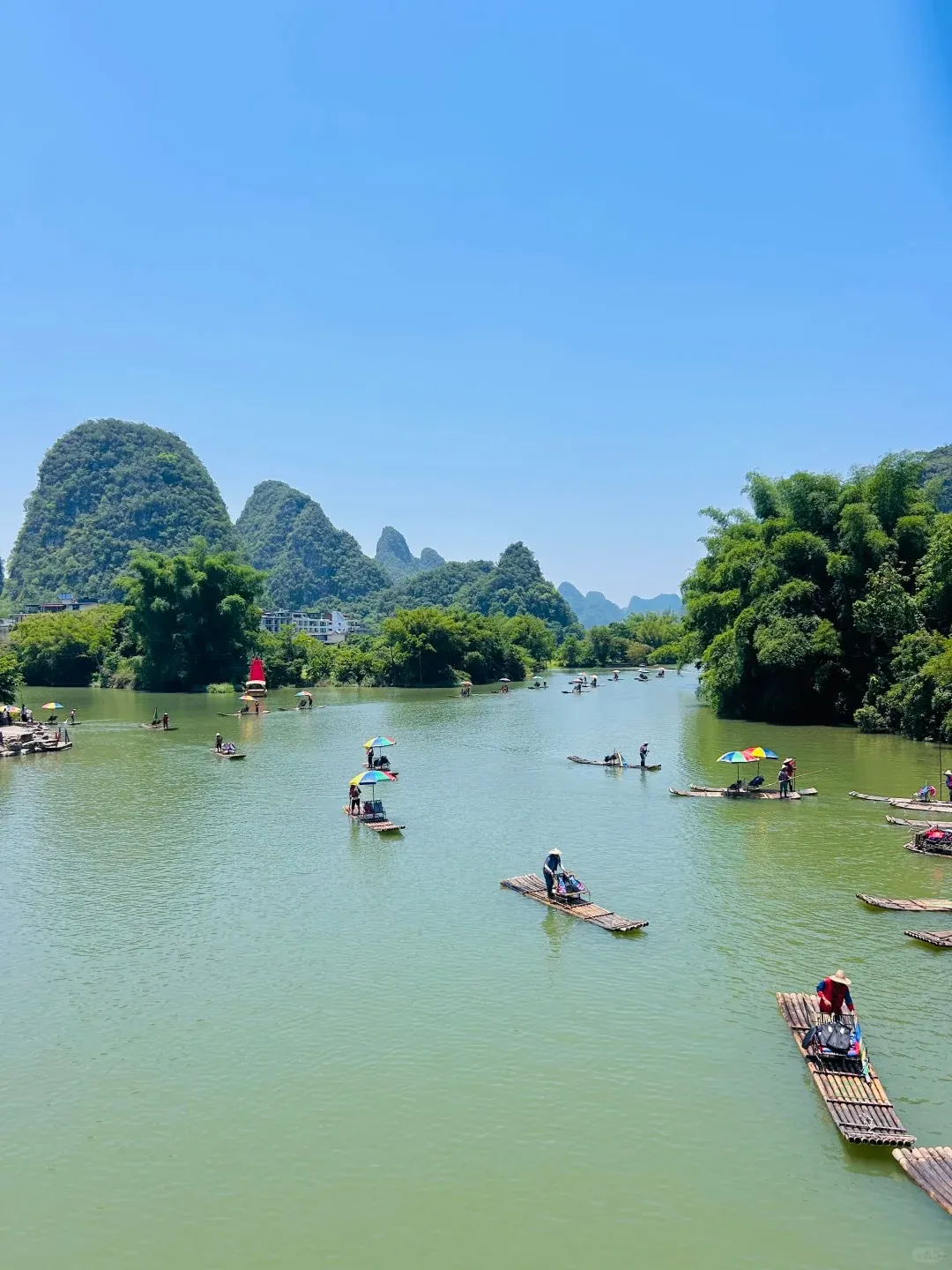 Many bamboo rafts with passengers and colorful umbrellas float on the green Yulong River, surrounded by lush, tree-covered karst mountains under a clear blue sky.