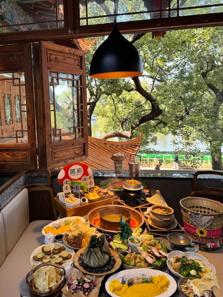 A large table laden with a grand feast, centered around a copper hotpot filled with orange broth. Surrounding the hotpot are numerous plates of Yunnanese dishes, including sliced mango, various salads, fried snacks, a wrapped dish, and a prominent mushroom-themed display with Chinese characters. The setting is a traditional wooden-paneled restaurant with large windows offering a scenic view of a lake, lush green trees, and distant people.