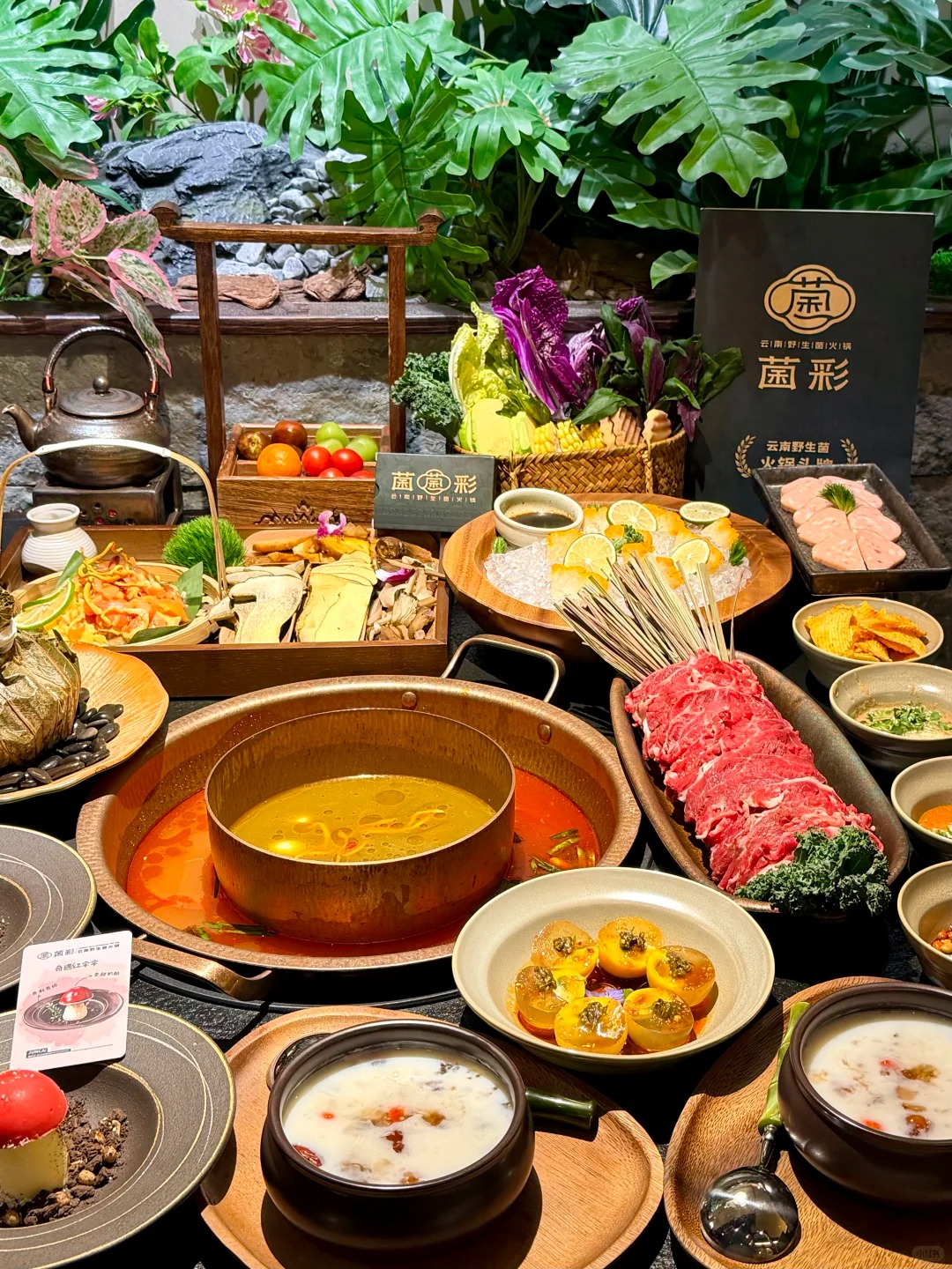 A large table is set with a traditional Chinese hotpot meal, featuring a copper split pot with two broths: one yellowish mushroom broth and one reddish spicy broth. Around the hotpot are various ingredients including plates of thinly sliced raw beef, fresh vegetables like purple cabbage and corn, a selection of dried and fresh wild mushrooms, sliced fish on ice, and baskets of fresh fruits. Several small bowls of sauces, two creamy desserts, and a mushroom-shaped cake are also visible. A menu for 'Yunnan Wild Mushroom Hotpot' stands in the background amidst lush green foliage.