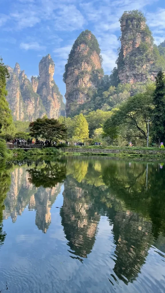 Towering sandstone pillars covered in green trees are reflected in a calm, dark green lake. On the far shore, a wooden walkway and paved path with numerous visitors are visible, framed by lush foliage under a partly cloudy blue sky.