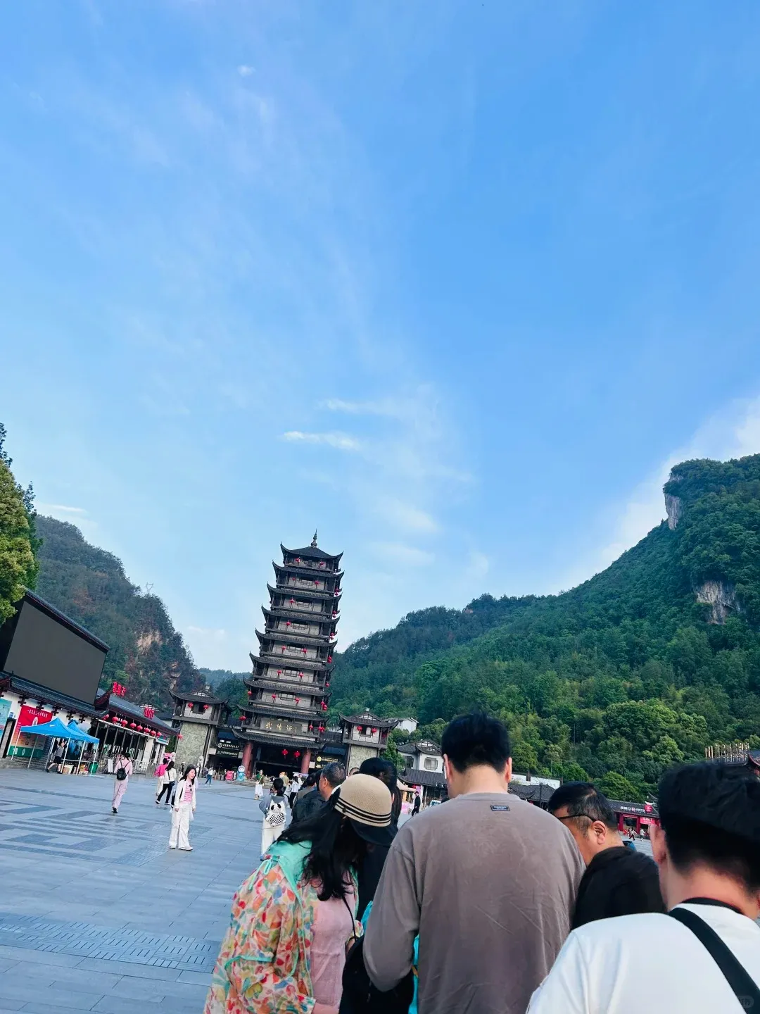 A large crowd of tourists gathers in an outdoor plaza, facing the multi-tiered traditional Chinese pagoda-style Wulingyuan East Gate in Zhangjiajie. The dark wooden gate features red accents and lanterns, nestled between lush green mountains under a bright blue sky.
