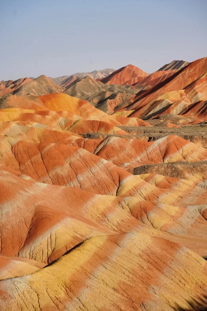 A wide shot of a mountainous landscape featuring numerous hills and peaks with distinct horizontal stripes of vibrant red, orange, yellow, and light grey rock. The arid terrain shows signs of erosion under a clear, pale blue sky.