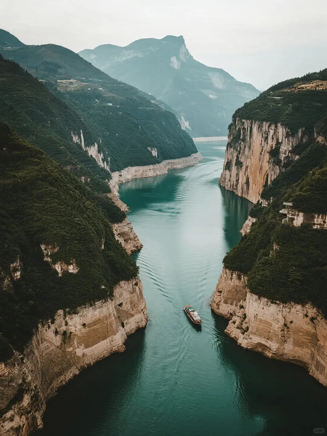 An aerial view showing a multi-deck cruise ship sailing on a wide, teal-colored river. The river is flanked on both sides by towering, steep cliffs covered in dark green vegetation and exposed light-colored rock formations. A gentle wake trails behind the ship as it proceeds into the distance under a bright, overcast sky.