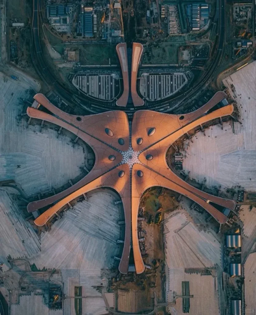 An aerial, overhead view of Beijing Daxing International Airport's main terminal building. The sprawling structure is brown and distinctively starfish-shaped with six radial piers emanating from a central hub, featuring several skylight domes. Surrounding the terminal are vast paved areas, active construction zones, railway tracks, and other airport support buildings, all bathed in warm, late afternoon sunlight casting long shadows.