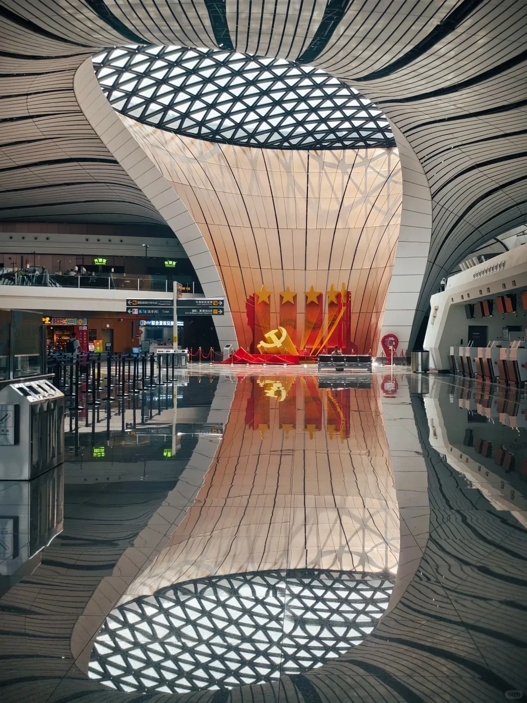 Interior of Beijing Daxing International Airport terminal featuring a large, open space with undulating white and grey ceiling panels, an oval skylight with a geometric grid, and a highly reflective dark floor. In the center, a tall, curved structure rises towards the skylight, with a red and gold display at its base featuring three large gold stars and a gold hammer and sickle-like emblem.