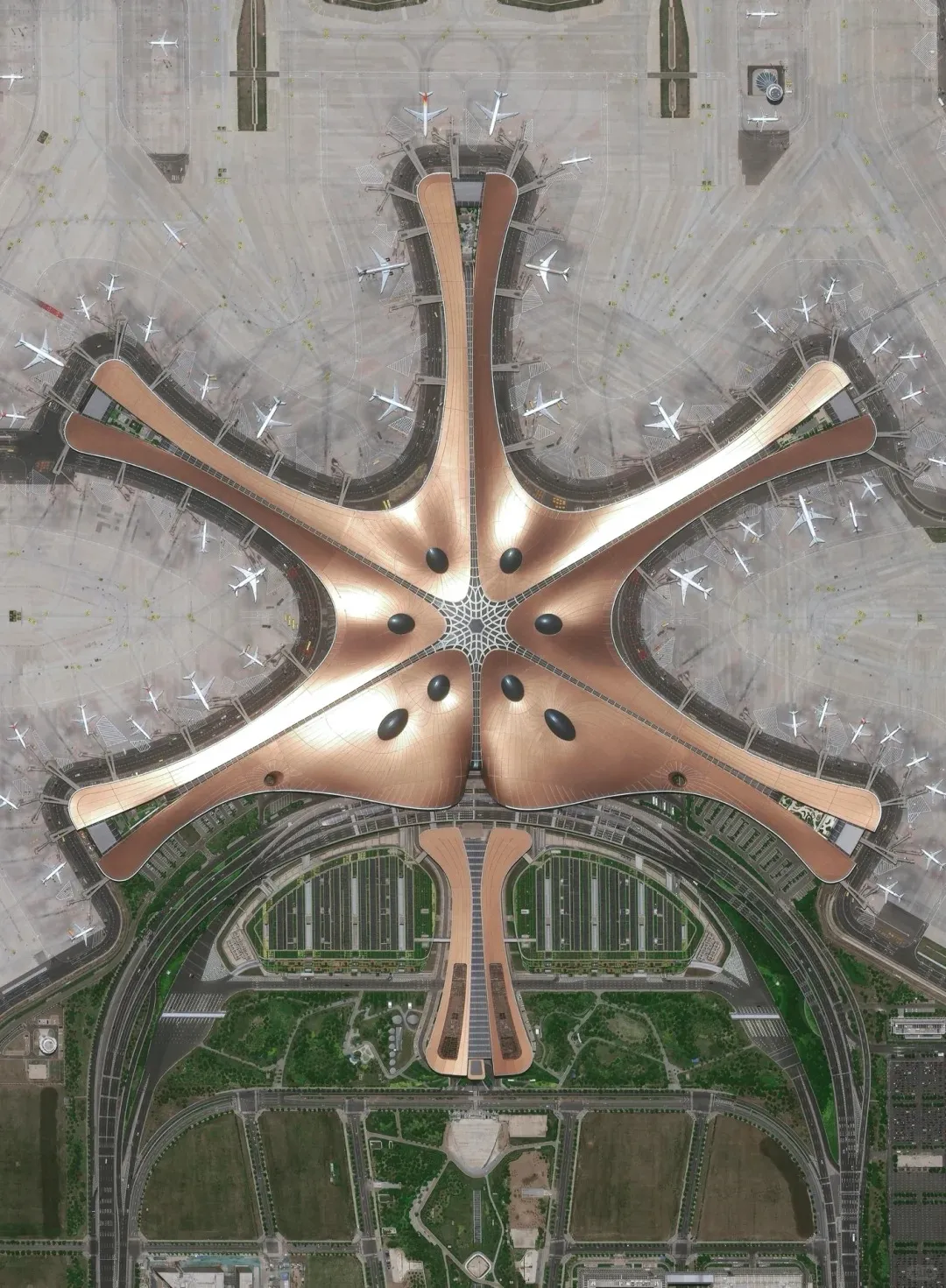 Aerial satellite view of Beijing Daxing International Airport's main terminal building. The sprawling structure is bronze-orange, shaped like a six-pronged starfish with a central hub. Numerous white passenger airplanes are docked along its long concourses, surrounded by vast concrete aprons, taxiways, and runways. Below the terminal, complex road networks and landscaped areas are visible.