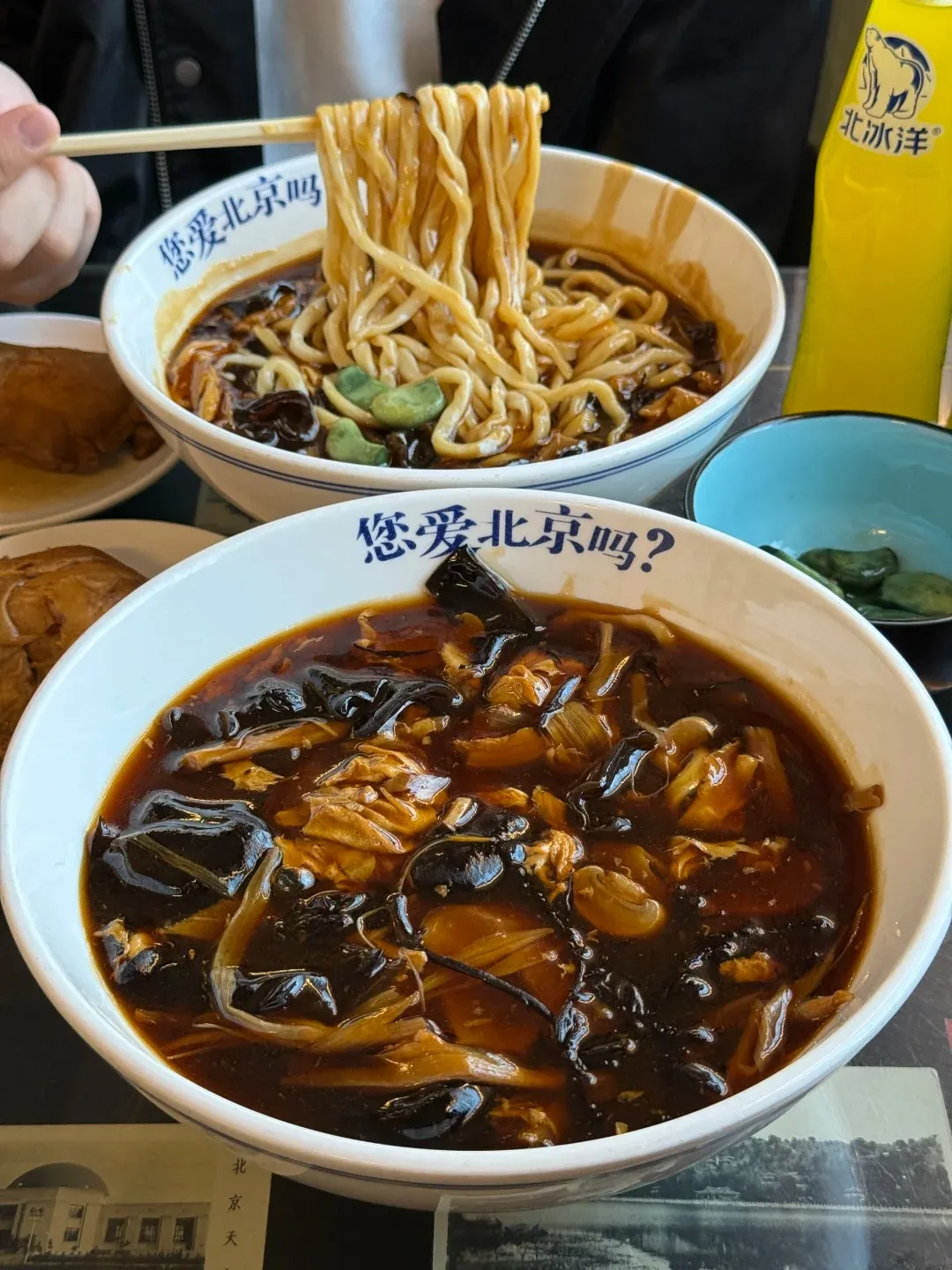 Two white bowls with blue rim and Chinese characters are filled with noodles and a dark brown sauce. A person's hand with chopsticks lifts a large serving of noodles from the top bowl, revealing ingredients like dark mushrooms, light shredded vegetables or meat, and green beans in the sauce. The bottom bowl is in the foreground, showing more of the dark brown sauce and ingredients. A yellow soda bottle, a small blue dish with pickles, and other side dishes are on the table.