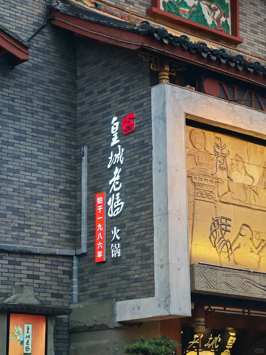 The exterior of Huangcheng Laoma hot pot restaurant in Chengdu, featuring a grey brick facade, a traditional tiled roof, and a prominent vertical illuminated sign displaying '皇城老妈火锅' (Huangcheng Laoma Hot Pot) and '始于一九八六年' (Founded in 1986). To the right, a large golden relief carving depicting traditional figures adorns the building.