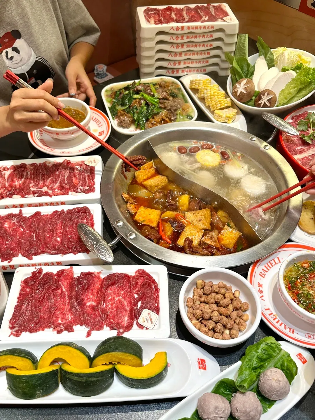 A large hot pot feast spread on a table, featuring a bubbling silver yin-yang hot pot with a fiery red spicy broth on one side and a clear, mild broth with corn and meatballs on the other. Surrounding the pot are multiple plates of thinly sliced raw beef, fresh vegetables including lettuce, cabbage, and shiitake mushrooms, corn on the cob, sliced raw pumpkin, a bowl of peanuts, and a dipping sauce. A person's hands hold red chopsticks, preparing to add ingredients to the hot pot. Stacks of white serving plates with raw beef and Chinese text are visible in the background.