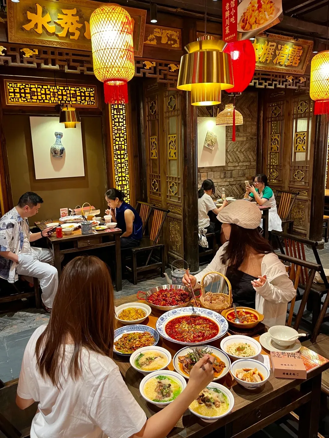 Interior of a traditional Chinese restaurant featuring dark wooden panels, ornate carvings, and hanging yellow and red lanterns. In the foreground, two women are seated at a wooden table overflowing with over ten colorful bowls of family-style Sichuan dishes, including prominent red chili dishes, various stir-fries, and noodle soups. Other diners are visible in the background.