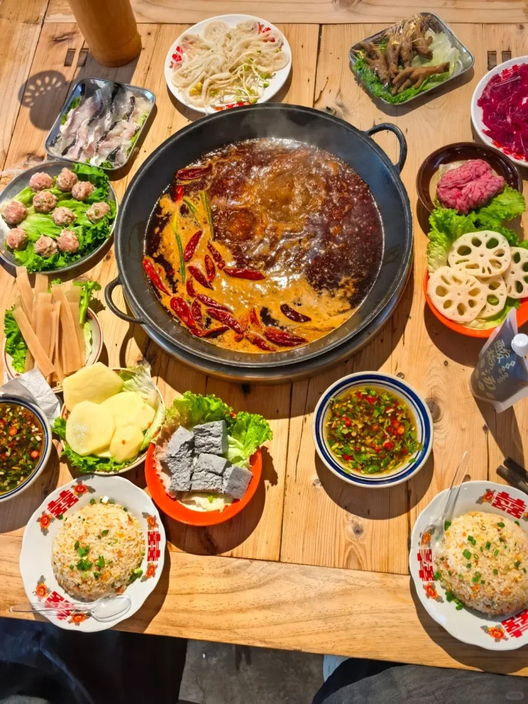 A vibrant overhead shot of a large, black Yin-Yang hot pot with bubbling red chili oil and clear broths, surrounded by numerous plates of raw ingredients on a wooden table. These include thinly sliced beef, fish fillets, tripe, meatballs, lotus root, potatoes, dark tofu or blood curd, seasoned chicken feet, two bowls of dipping sauce, and two plates of fried rice.
