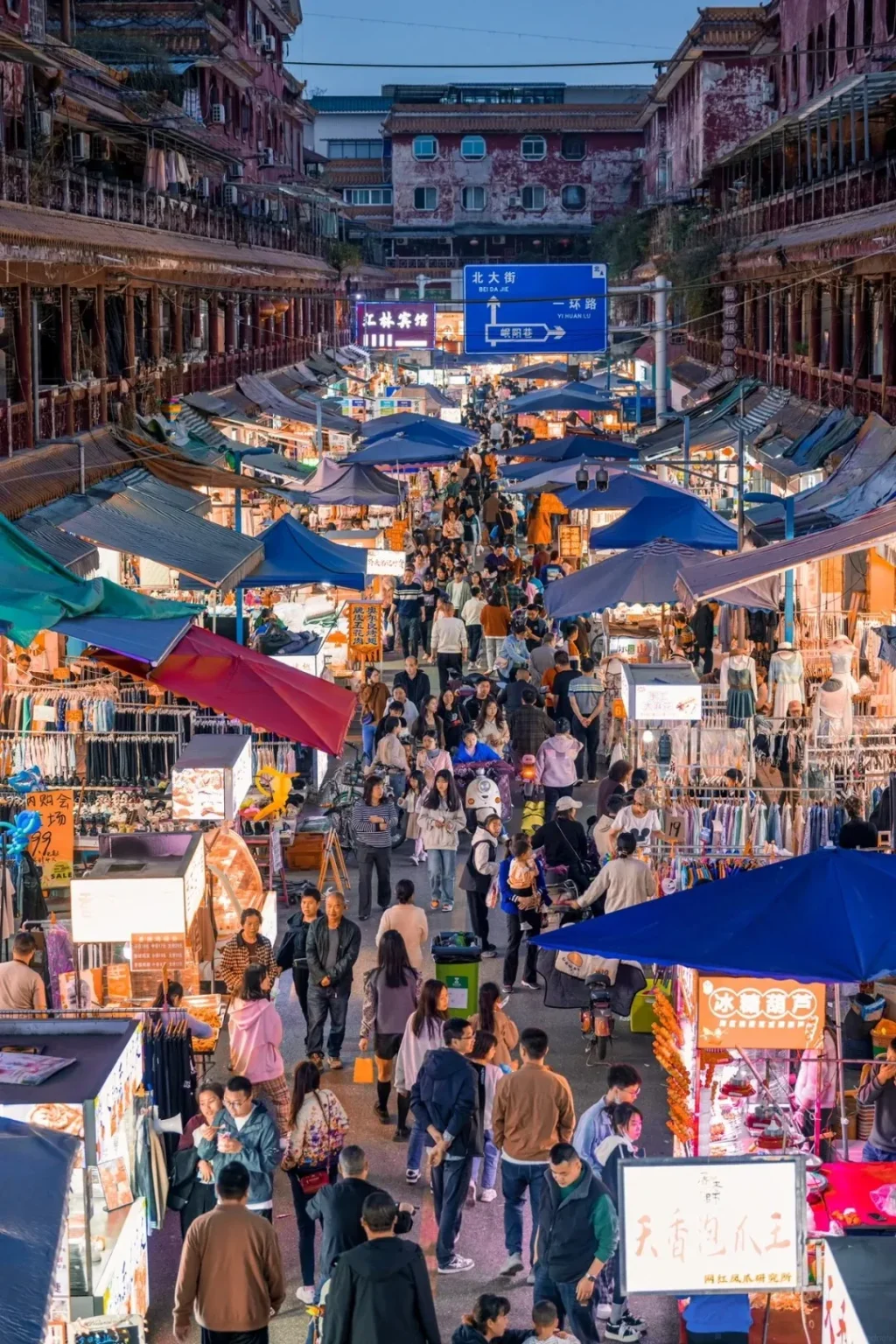 A bustling outdoor night market in a Chinese city, viewed from a slightly elevated perspective. Numerous people are walking along a wide street lined with dozens of stalls covered by blue, red, and various colored tarps. Racks of clothing, merchandise, and illuminated food displays are visible at the stalls. Traditional red-roofed buildings with weathered facades line both sides of the market street in the background, with blue street signs displaying Chinese characters visible above the crowd.