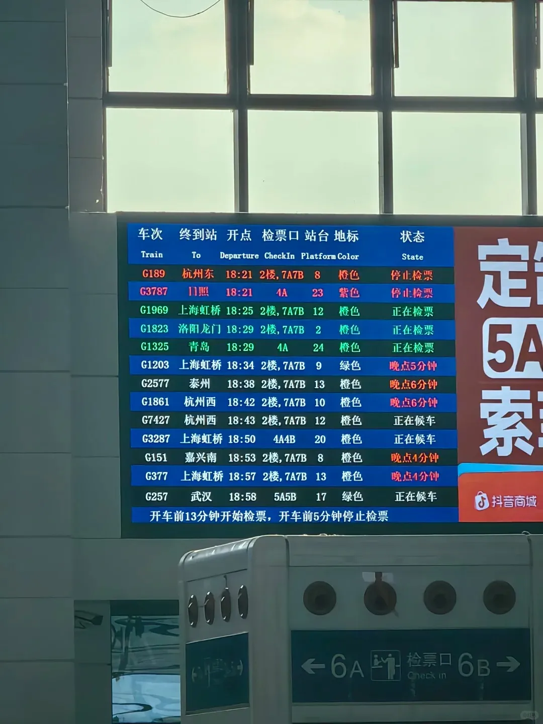 A digital departure board at a busy Chinese high-speed railway station, displaying train information in both Chinese and English. Columns include Train, To, Departure, CheckIn, Platform, Color, and State. Multiple train entries are visible with numbers, destinations like Hangzhou East and Shanghai Hongqiao, departure times, gate numbers, and statuses such as 'Stopped check-in,' 'Checking in,' 'Waiting,' and 'Delayed 6 minutes.' Below the main list, text in Chinese and English explains check-in timings. A grey check-in gate with '检票口 Check in' and arrows for gates 6A and 6B is partially visible at the bottom.