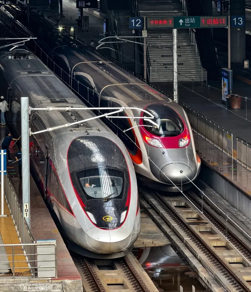Two modern high-speed trains, one silver with red accents and a black front, and another silver with red and pink accents, are side-by-side on parallel tracks at a train station. People are visible on the platform near the front of the silver train. Overhead signs indicate platform numbers and directions to exits and transfers.