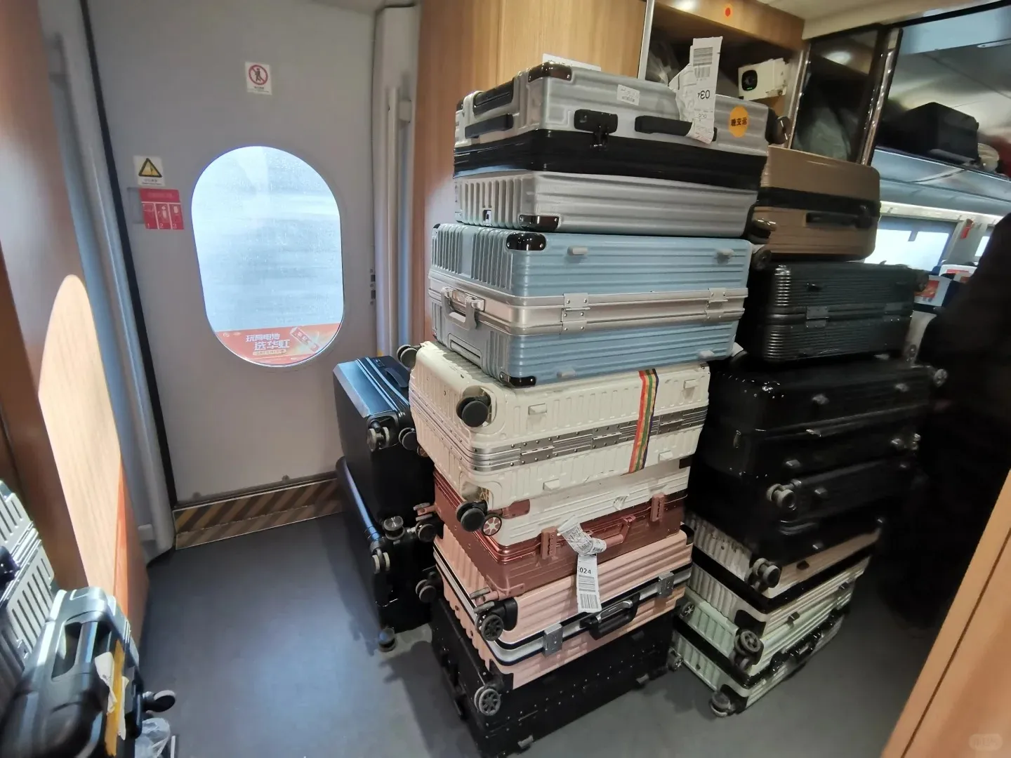 Inside a modern train compartment, multiple stacks of colorful hard-shell suitcases are piled high in a narrow storage area next to a light gray door with an oval window. The luggage includes silver, light blue, white, pink, black, brown, and light green suitcases, some with paper travel tags.