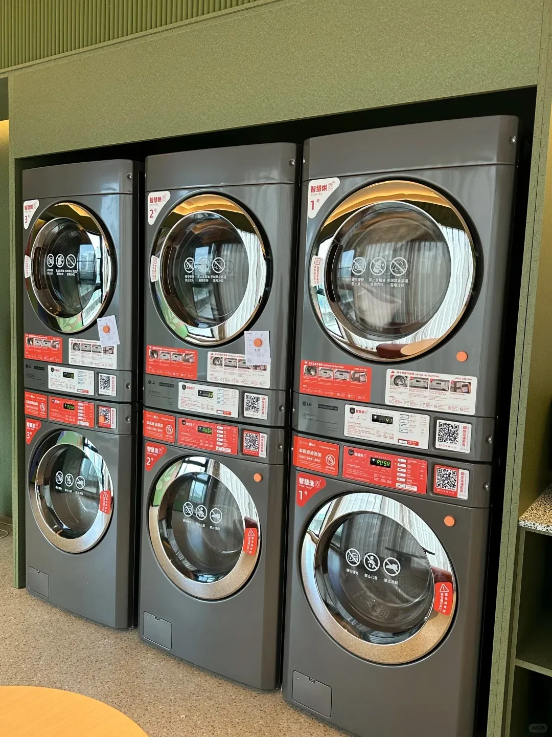 Three sets of dark grey stacked LG brand washing machines and dryers are arranged side-by-side against a muted green paneled wall in a modern laundry room. Each unit features a prominent circular glass door, and red instruction panels with Chinese text and QR codes are visible between the stacked appliances.