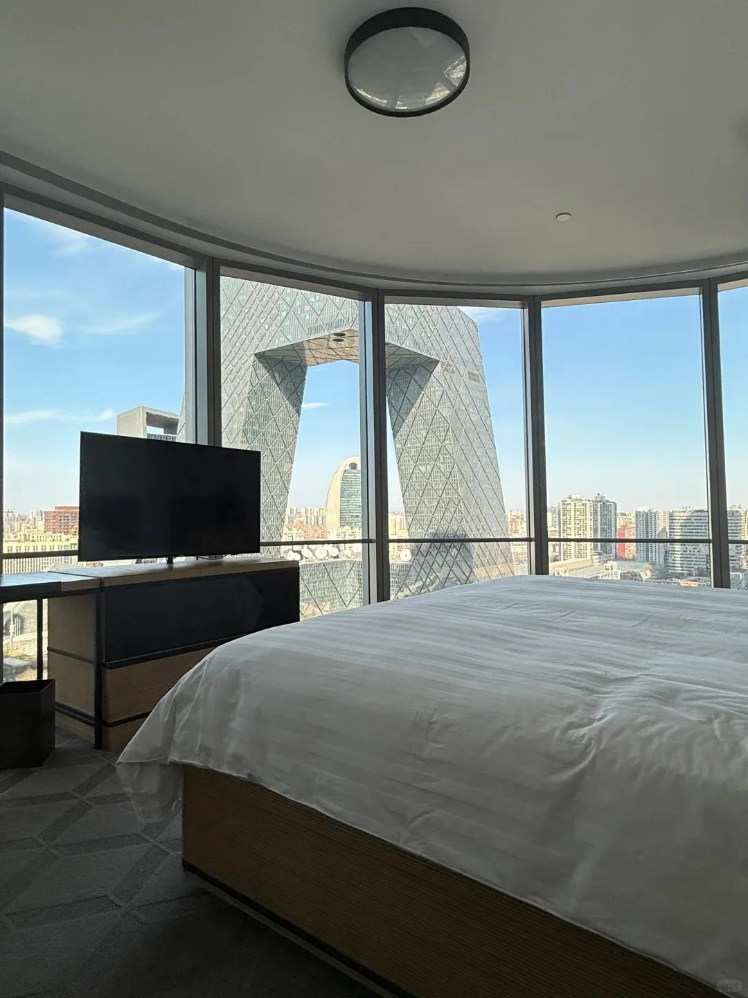 A modern hotel room with a large white bed and a black flat-screen TV, offering a panoramic view of the Beijing cityscape through floor-to-ceiling windows, with the distinctive CCTV Headquarters building prominently visible under a blue sky.