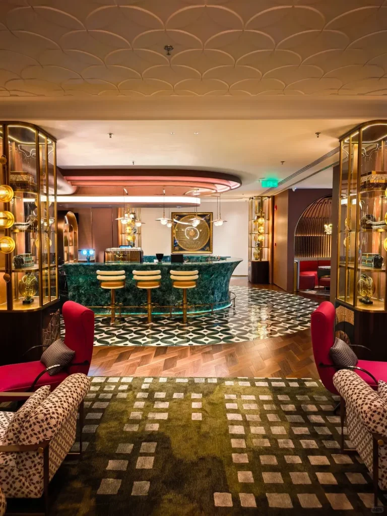 Luxurious hotel lobby bar featuring a curved emerald green marble counter with three cream and gold bar stools. The floor transitions from dark geometric tiles to herringbone wood, with an olive green geometric rug in the foreground. Red lounge chairs and patterned armchairs are visible. Ornate gold display cases flank the sides, and a large circular artwork hangs above the bar.