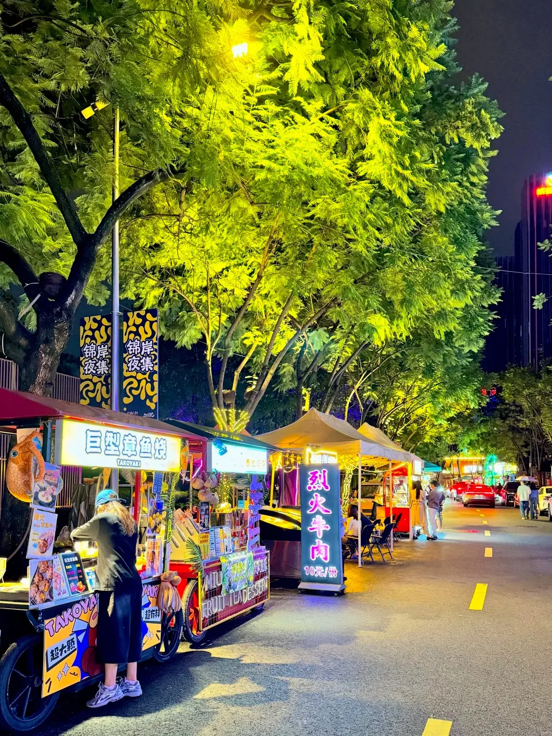A person stands at a food cart in a bustling night market in China, illuminated by bright lights from the stalls and trees. The cart displays various food items and menus with Chinese characters. The person is partially obscured by the cart and appears to be interacting with it. Other food stalls and tents are visible further down the street, along with people and cars. The street is paved with yellow lane markings.