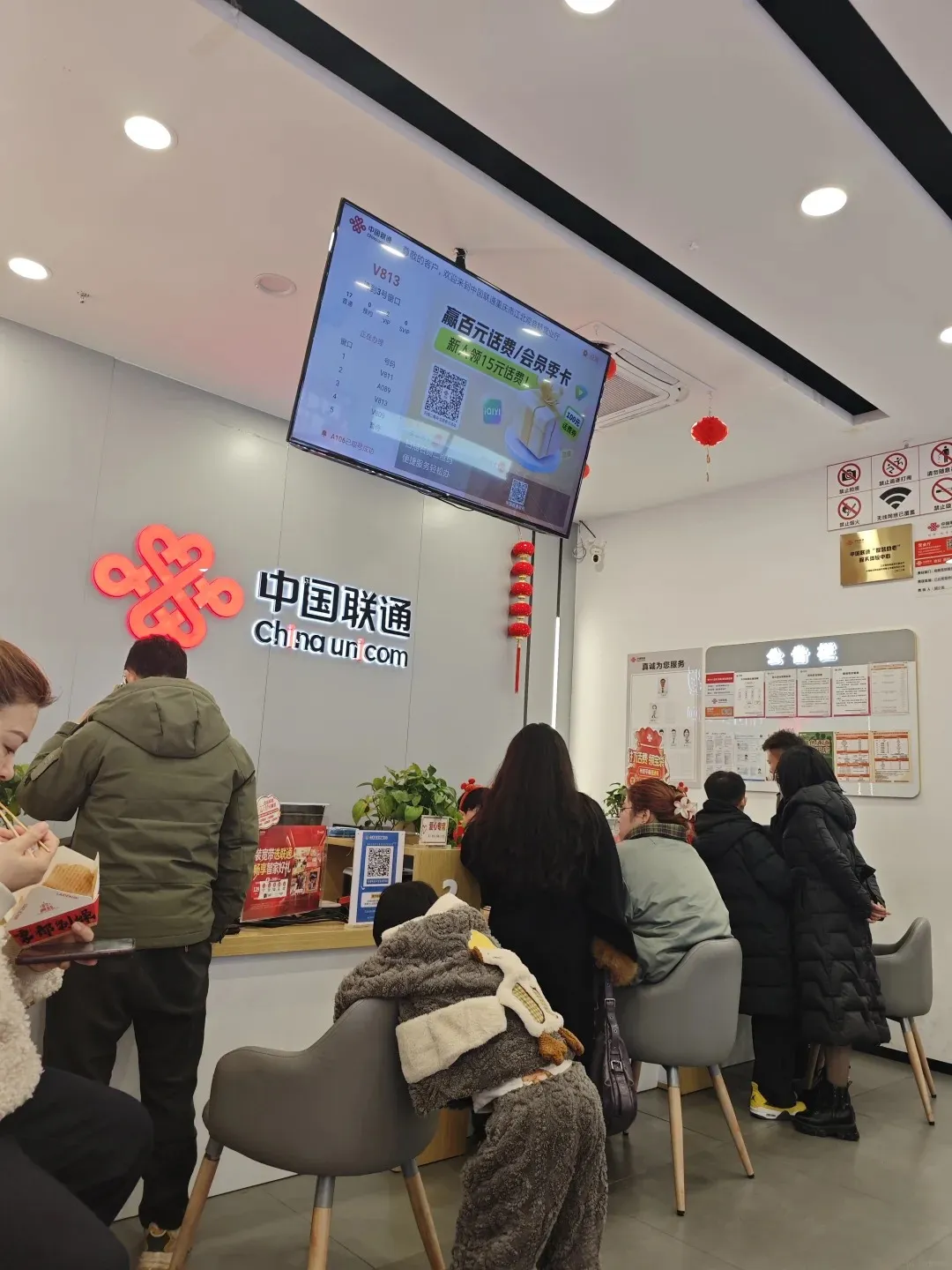 A busy China Unicom store interior with multiple customers waiting at service counters. A large digital screen displays queue numbers and advertisements above the main desk. People are standing and sitting on modern grey chairs, while red lanterns hang from the ceiling. The '中国联通 chinauni.com' logo is visible on the back wall, and a 'no smoking' sign is on the right.