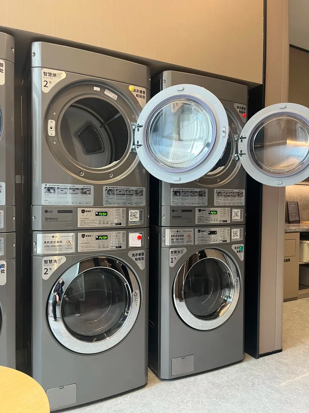 Two pairs of stacked grey LG commercial washer and dryer machines in a modern self-service laundry room. The top dryers on both the left and right units have their doors open, revealing their interiors. Each machine features a front-loading door and control panels with Chinese text and QR codes.