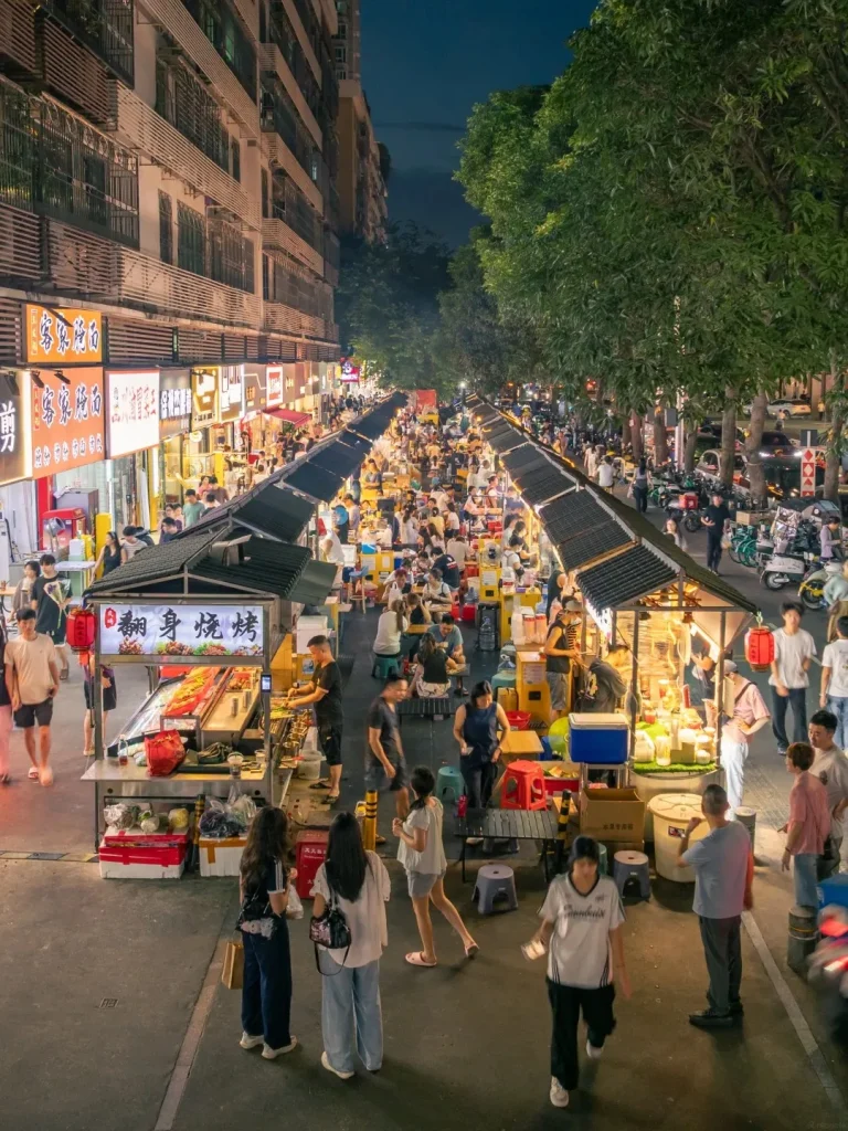 An aerial view of a bustling Chinese street food market at dusk, with numerous food stalls lining a street. People are gathered around the stalls, ordering and eating. The stalls are lit, displaying various food items and signs in Chinese. Trees line one side of the street, and apartment buildings rise on the other. The overall scene depicts a vibrant and crowded marketplace.
