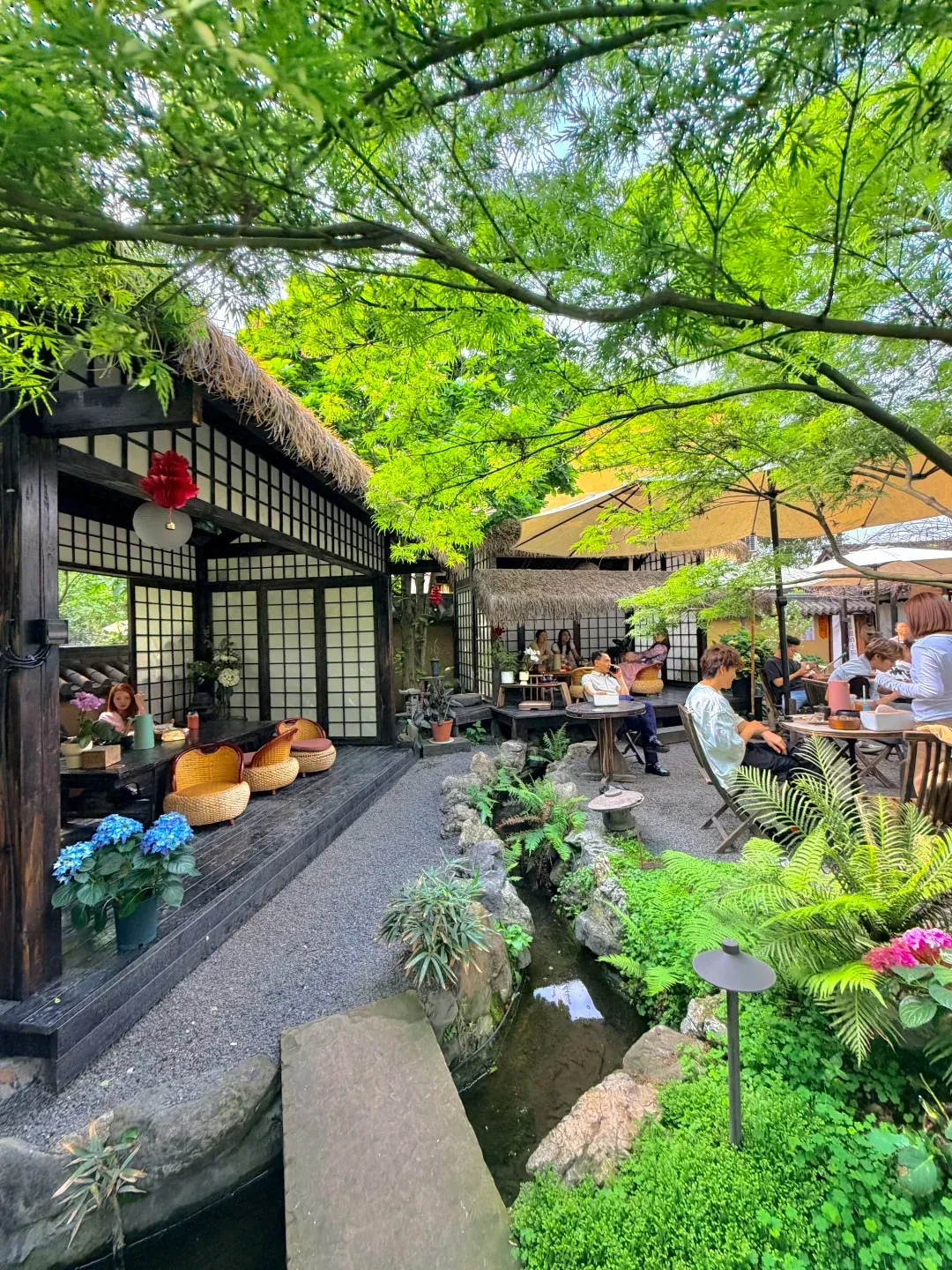 A traditional Chinese teahouse with a thatched roof, dark wooden beams, and sliding shoji screen doors. In the foreground, several people are seated at tables and on bamboo chairs in a tranquil courtyard. A small, winding stream flows through the garden area, surrounded by lush green plants and rocks. Blue hydrangeas in a pot and a red paper lantern hanging from the eaves add pops of color. Sunlight filters through the leafy green trees overhead.
