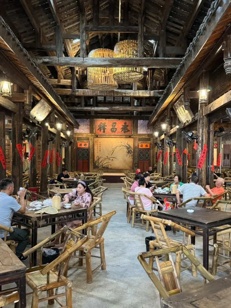 A wide shot of a traditional Chinese teahouse interior with high wooden beamed ceilings, hanging woven bamboo light fixtures, and numerous tables and chairs made of bamboo. People are seated at several tables, some dining and some drinking tea. Red banners with Chinese characters hang vertically from wooden pillars. A large mural depicting a landscape scene with birds is visible on the back wall.
