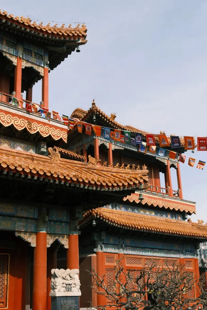 Multi-tiered traditional Chinese temple buildings feature orange tiled roofs, red pillars, intricately painted eaves, and colorful prayer flags strung between levels. A stone sculpture is visible near a red pillar, with a bare tree branch in the foreground.