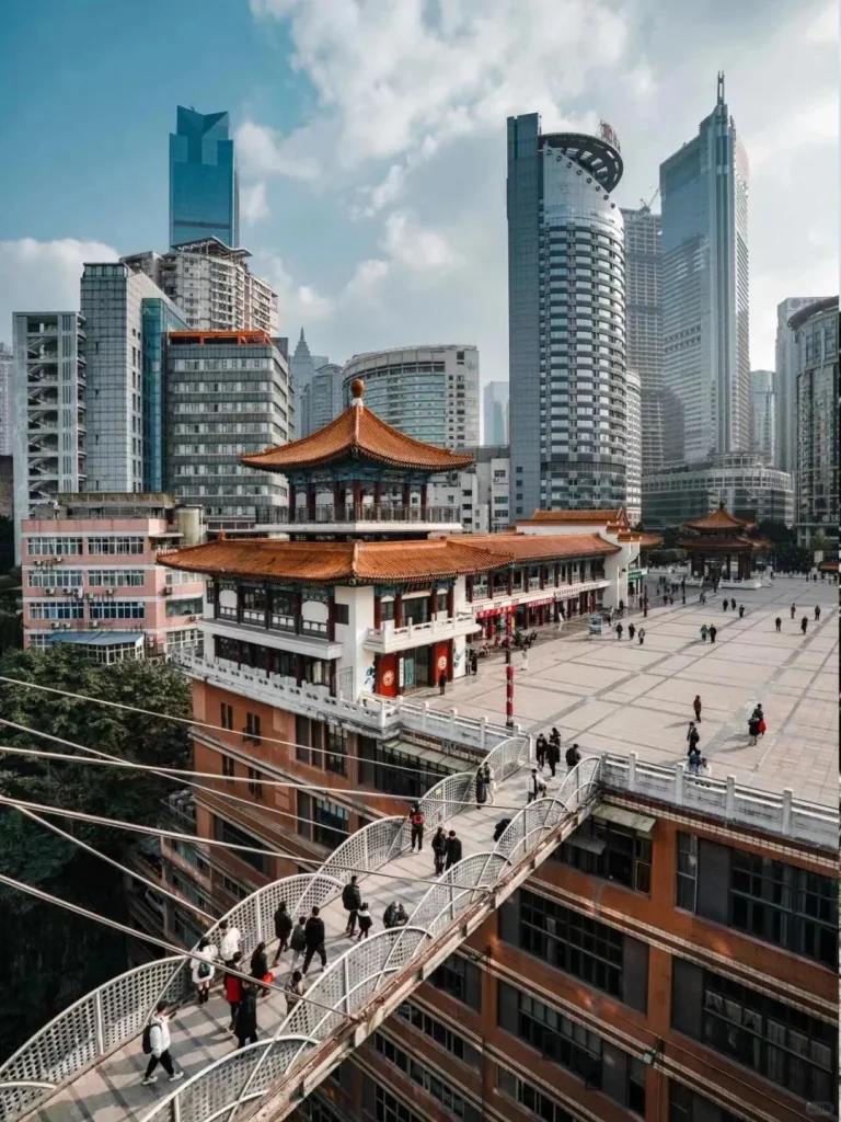 An aerial perspective of Chongqing's dynamic cityscape. In the foreground, a curved, white pedestrian bridge, bustling with people, spans between buildings. The bridge leads to a large, open public plaza next to a traditional Chinese building featuring an ornate, orange-tiled, multi-tiered roof. Tall, modern glass skyscrapers dominate the background under a partly cloudy sky, illustrating the city's complex vertical topography and architectural blend.