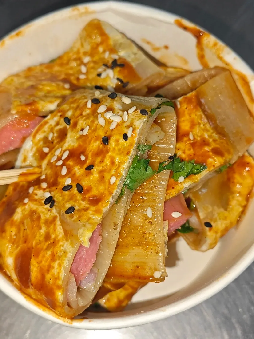 A close-up shot of a Jianbing, a Chinese breakfast crepe, folded and sliced in a white paper bowl. The crepe is filled with pink sausage and a cooked egg, and drizzled with a reddish-brown sauce. It is sprinkled with white and black sesame seeds and fresh green cilantro. A chopstick is inserted into the crepe.