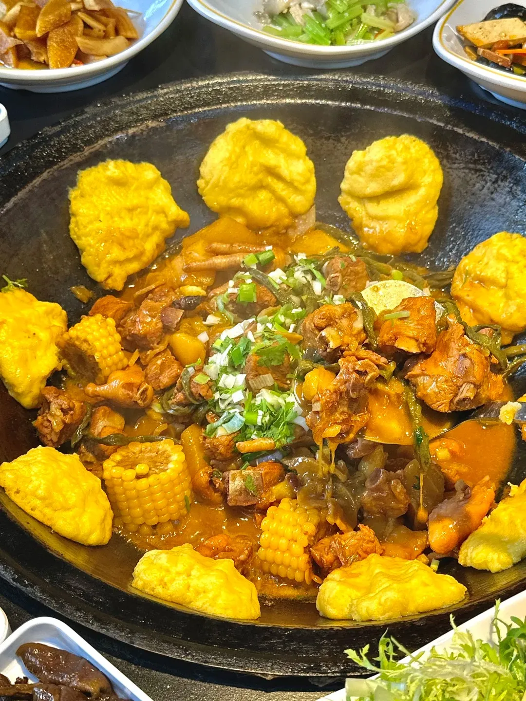A large, dark, round cast-iron wok filled with a hearty stew. The stew contains visible pieces of meat, likely ribs, along with chunks of corn on the cob, potatoes, and green onions. Surrounding the stew are several golden-yellow, fluffy cornmeal buns. The dish is steaming, indicating it is hot. In the background, out of focus, are small white bowls containing other food items.