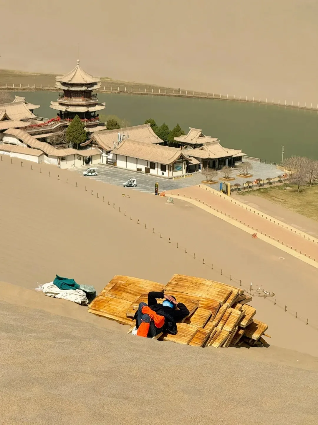 A high-angle photo from a sand dune showing a person resting on wooden boards in the foreground. Below, a cluster of traditional Chinese buildings featuring curved roofs and a multi-story pagoda is situated next to a long, green body of water. Distant golden sand dunes stretch across the background.