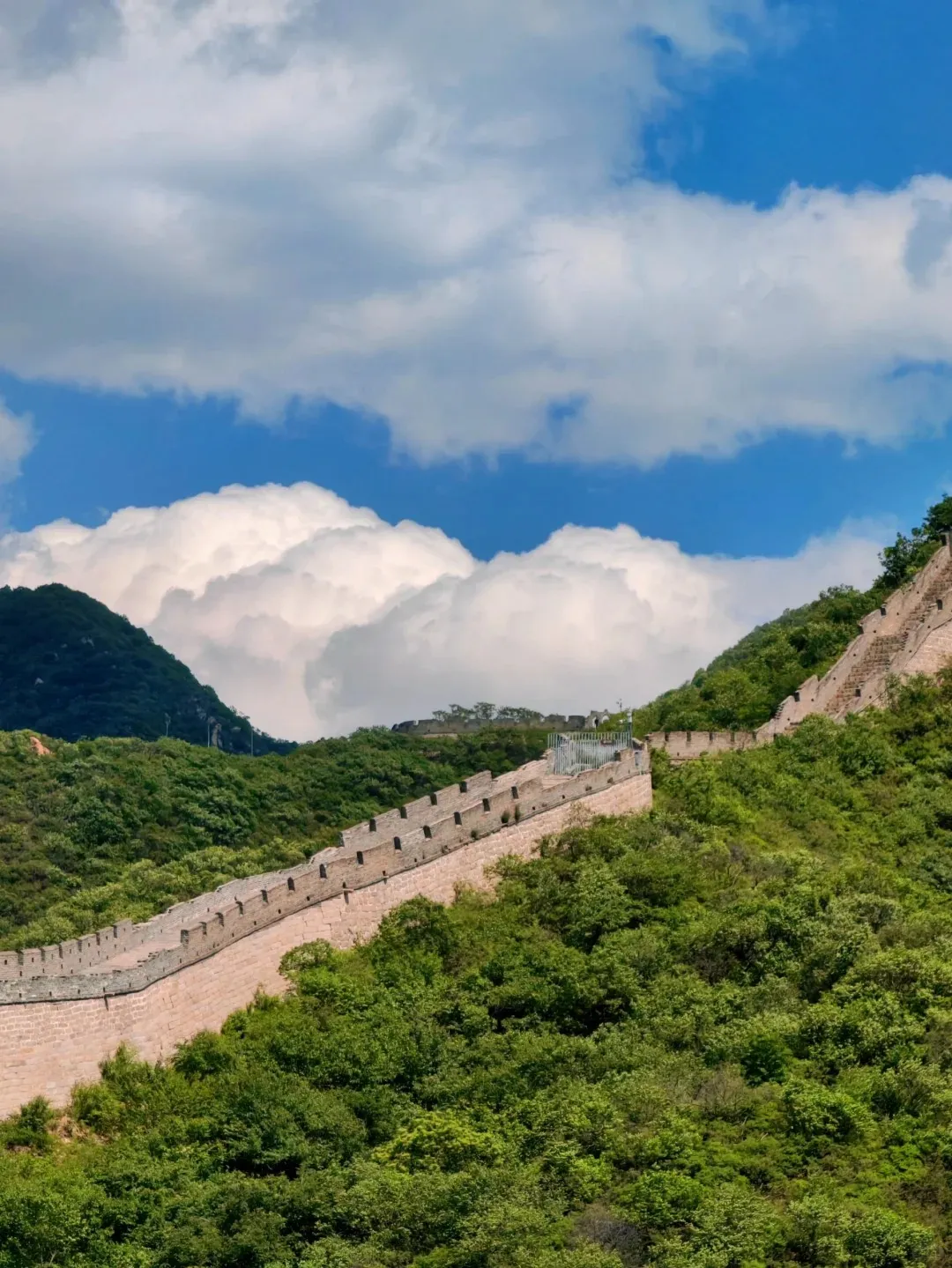A long, light-colored stone section of the Great Wall of China winding up and down lush green, tree-covered mountains. The wall features several watchtowers and battlements, all set against a bright blue sky filled with large white cumulus clouds.