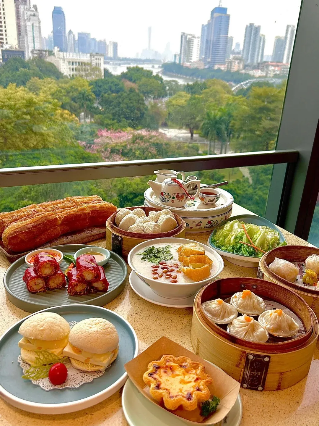 A table filled with a diverse dim sum spread, including long fried dough sticks, red rice noodle rolls with shrimp, pineapple bun sandwiches, congee with fried dough, an egg tart, steamed greens, and various dumplings in bamboo steamers, alongside a traditional Chinese teapot set. In the background, a blurred cityscape of Guangzhou with skyscrapers, a river, and green trees is seen through a window.