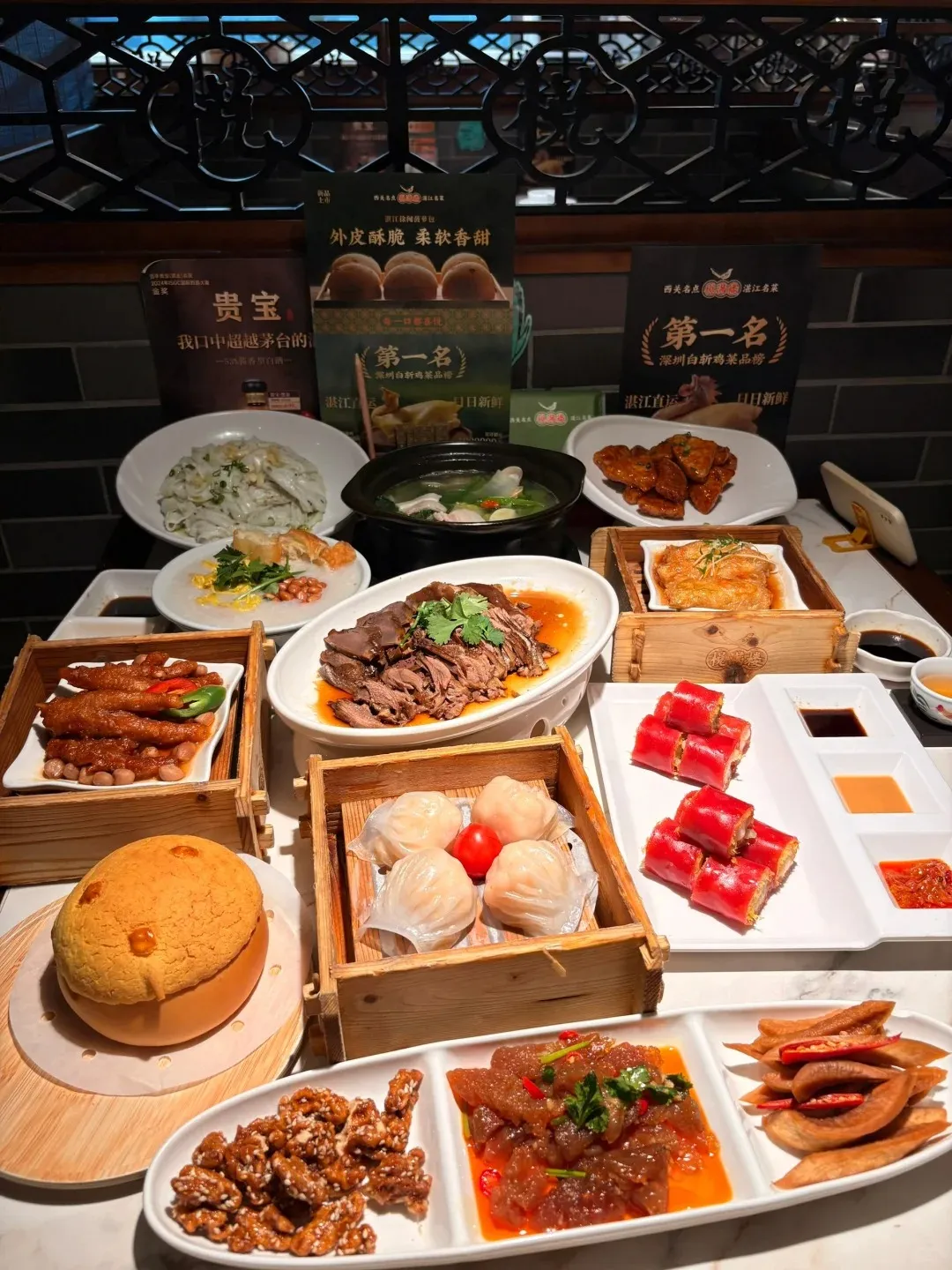 A wide shot of a restaurant table laden with a diverse spread of Cantonese dim sum and dishes. Prominently featured are steamed shrimp dumplings in a wooden box, red rice noodle rolls with dipping sauces, a large pineapple bun, braised chicken feet, sliced braised meat, a soup pot, candied walnuts, a spicy jelly dish, and pickled vegetables. Promotional posters for food products are visible in the background.