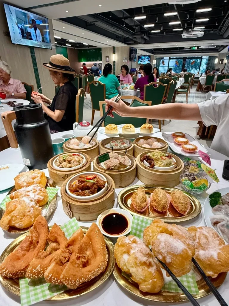 A large white dining table at a bustling Chinese dim sum restaurant is covered with a wide variety of dishes. Bamboo steamers hold different types of steamed dumplings, while plates display golden-brown fried sesame-coated pastries, sugar-dusted fried dough, taro puffs, and small bowls contain chicken feet and colorful jelly. A black tea thermos, a small white teapot, and a bowl of soy sauce are also on the table. In the foreground, a person's hand uses chopsticks to pick a dumpling from a steamer basket. Other diners are seated at tables in the background with a television screen mounted on the wall.