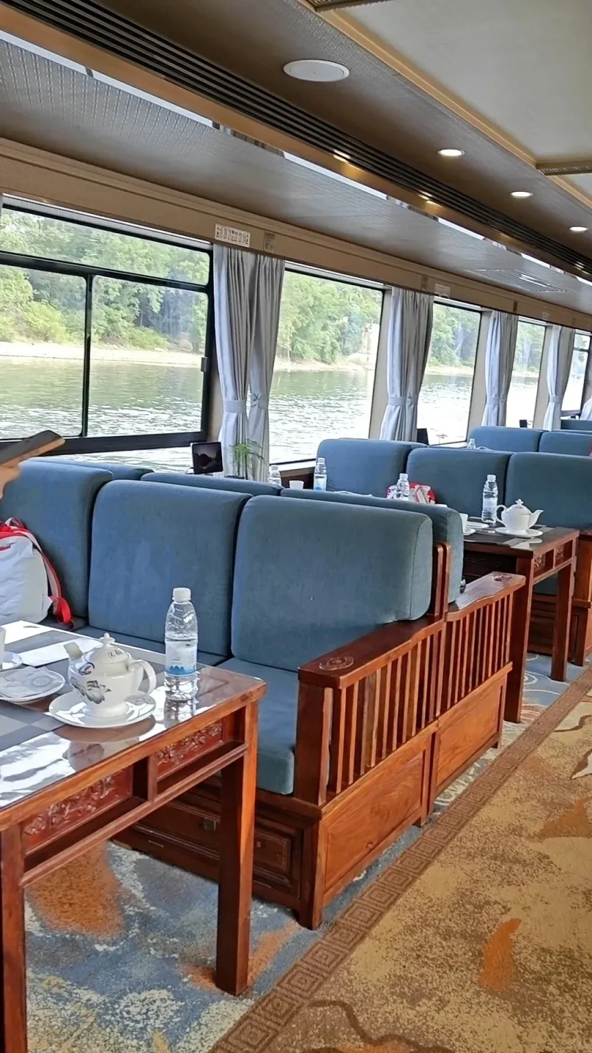 Interior view of a Li River cruise boat showing a comfortable lounge area. Rows of blue cushioned seats and ornate wooden tables line large windows, which overlook the river and lush green banks. Teapots and bottled water are on the tables.